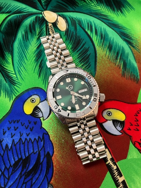 Islander wrist watch with green dial stainless steel band with the time set at 6:22, Thursday, the 23rd, sitting on top of a Hawaiian shirt fabric. Fabric is green back ground with a palm tree and two tropical birds. A blue bird to the left of the watch and a red bird on the right.